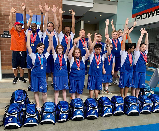 Two dozen people in blue shirts and blue shorts with olympic medals around their necks stretch their arms into the sky while smiling